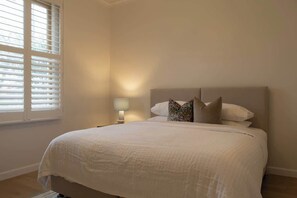 A bedroom with a large bed, a nightstand with a lamp, and a window with blinds. - Luxe Heritage Retreat - Sleeps 9 | CBD Edge (Bendigo)