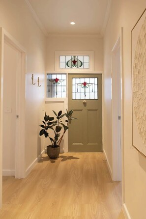A hallway with wooden flooring, a potted plant, a door with a glass panel, and a wall-mounted light fixture. - Luxe Heritage Retreat - Sleeps 9 | CBD Edge (Bendigo)