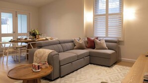 Ein Wohnzimmer mit einer Eckcouch, einem Esstisch mit Stühlen und einem großen Fenster mit Jalousien.