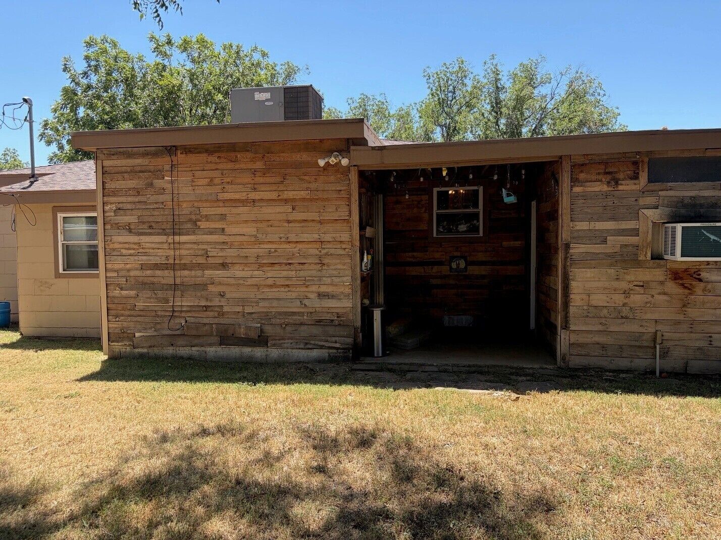 A rustic wooden building with a single door and a small window, surrounded by dry grass and trees.
