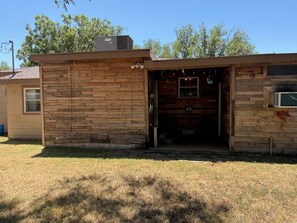 A rustic wooden building with a single door and a small window, surrounded by dry grass and trees.