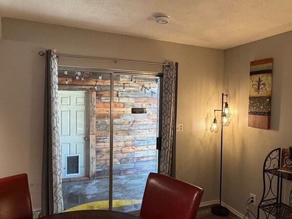 Una habitación con puerta de vidrio corredera que da a un balcón, una mesa de comedor con sillas, una lámpara fija en la pared y un pequeño estante con objetos.