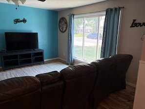 A living room with a flat-screen TV on a stand, a brown leather couch, a blue wall with the word 'Family' on it, and a window with curtains.