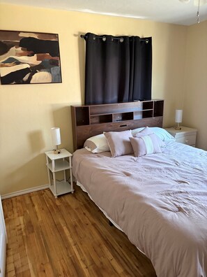 Un dormitorio con piso de madera, una cama con ropa blanca, una mesita de noche con una lámpara y un cuadro en la pared.