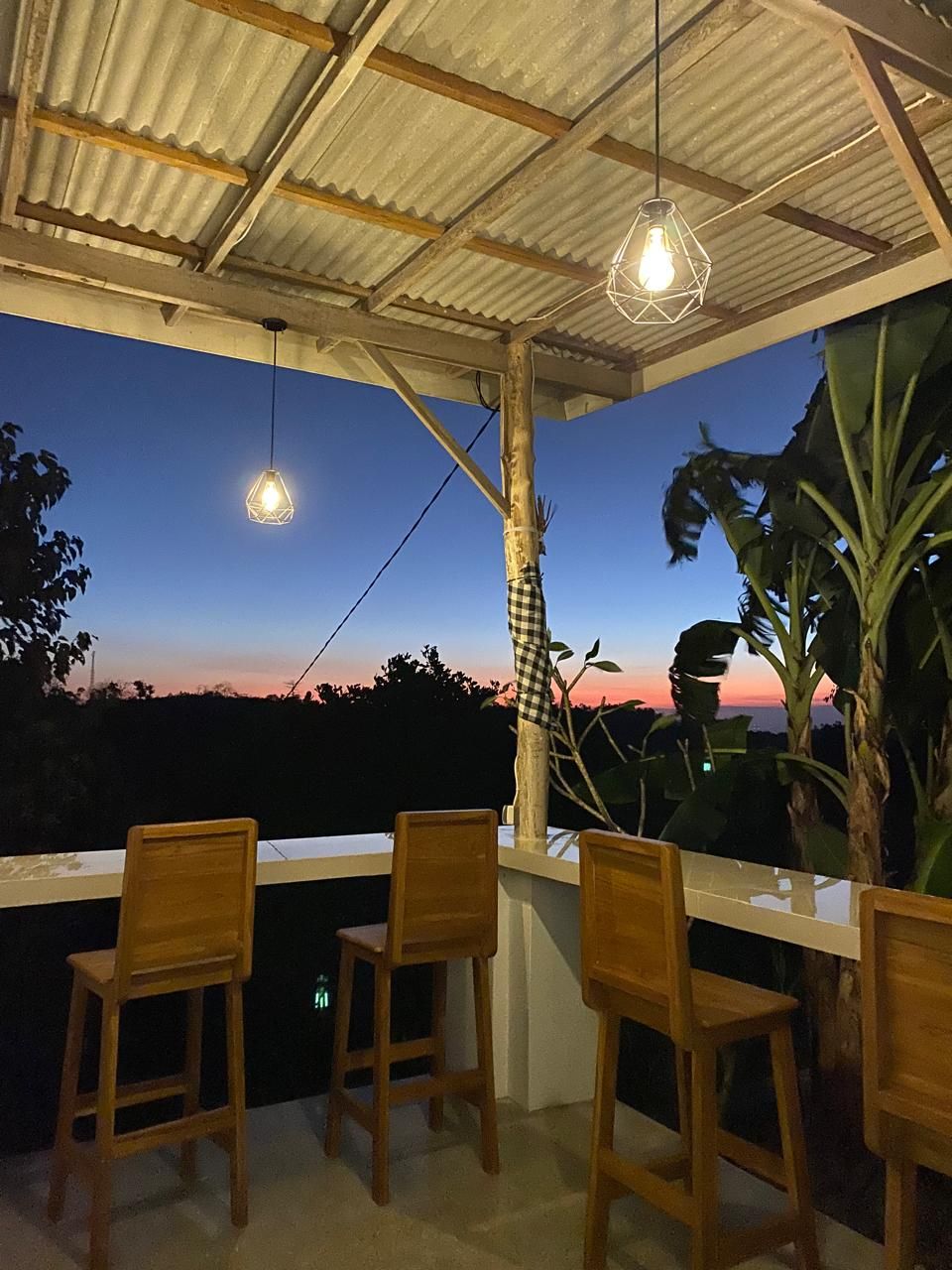 An outdoor dining area with wooden chairs and a white table, under a corrugated metal roof with hanging light fixtures.