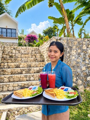 A person holding a tray with two plates of food and two drinks, standing in front of a stone wall with a house and tropical plants in the background. - Penida Cabins (Penida Island)