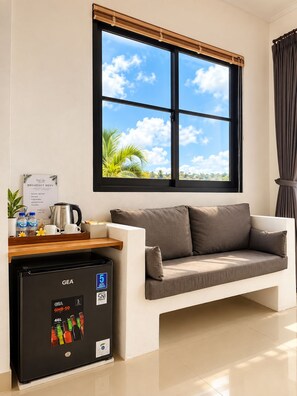 Una habitación moderna con un ventanal amplio, un sofá, una mesita de centro, un refrigerador GEA y un cartel con el menú de desayuno.
