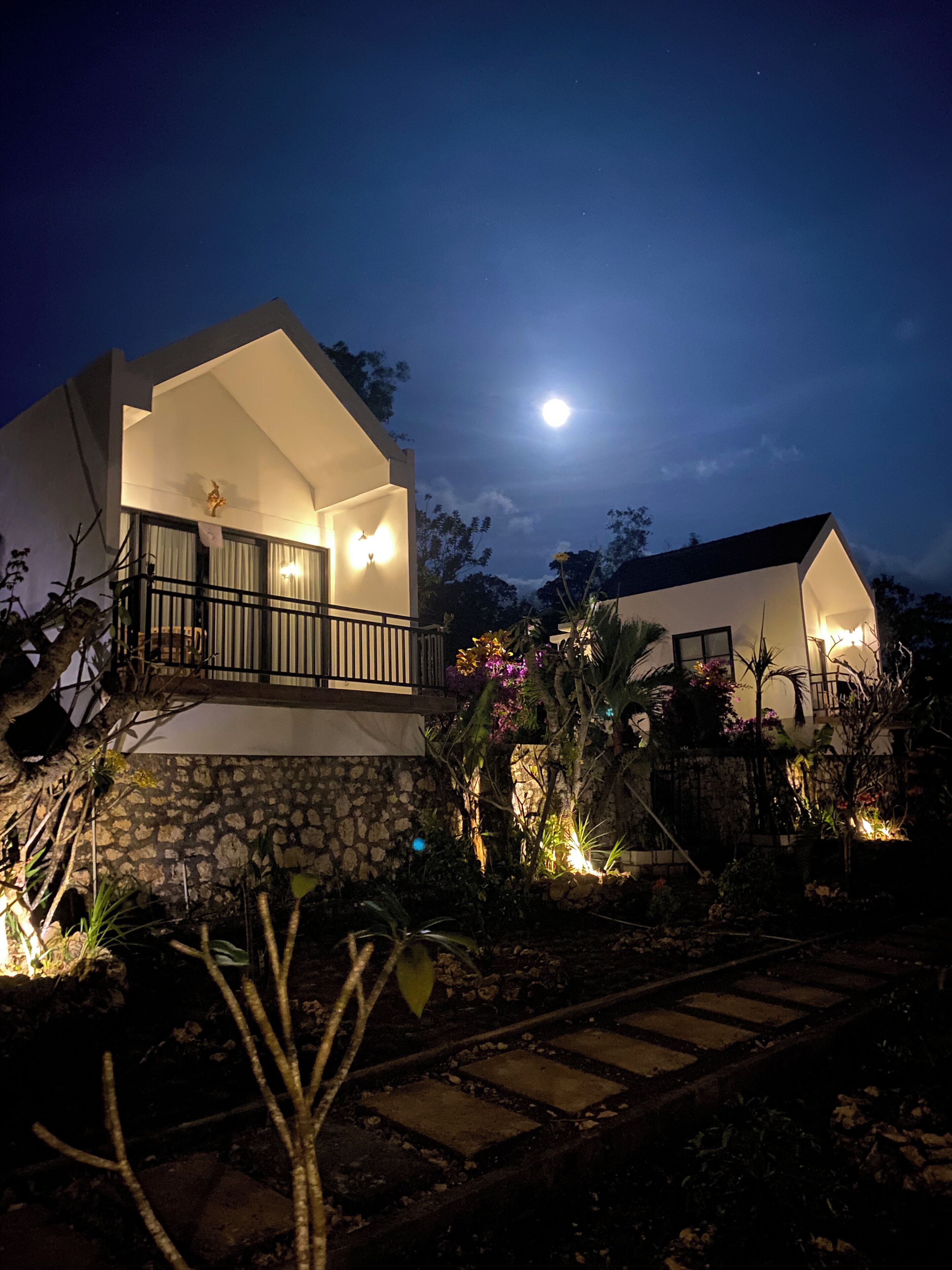 Una casa moderna con balcón, base de piedra y un jardín con plantas y un camino.