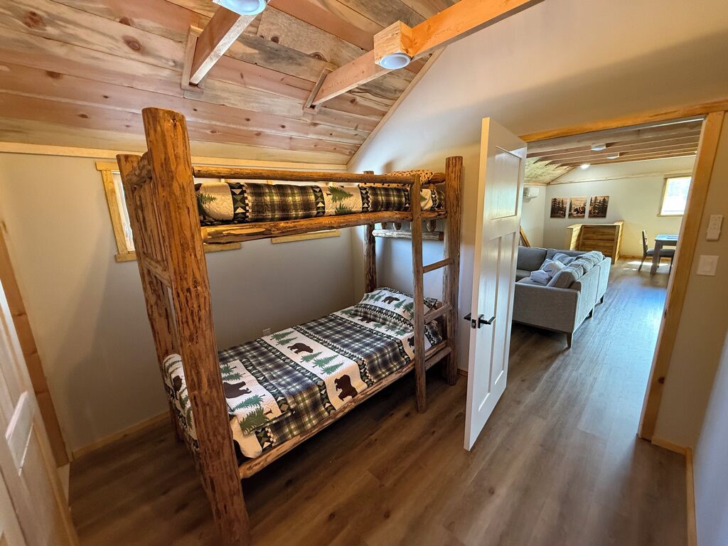 Una cama litera con una colcha de estampado escocés, techo de madera y una puerta que da a una sala de estar con sofá.