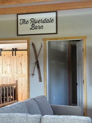 Ein Zimmer mit einem Schild, auf dem 'The Riverdale Barn' steht, einer Holztür, einem Kleiderständer mit Skiern und einem grauen Sofa.