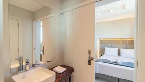 Ein Hotelzimmer mit einem großen Spiegel, einem Waschbecken, einer Holzkommode, einem Bett mit weißen Leinen und einem Kopfteil in heller Holzoptik.