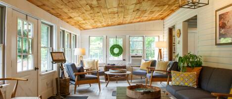 A cozy living room with wooden ceiling, large windows, and a fireplace.