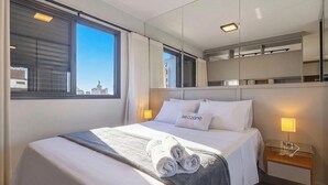 Een hotelkamer met een bed, nachtkastje, lamp en uitzicht op de stad.