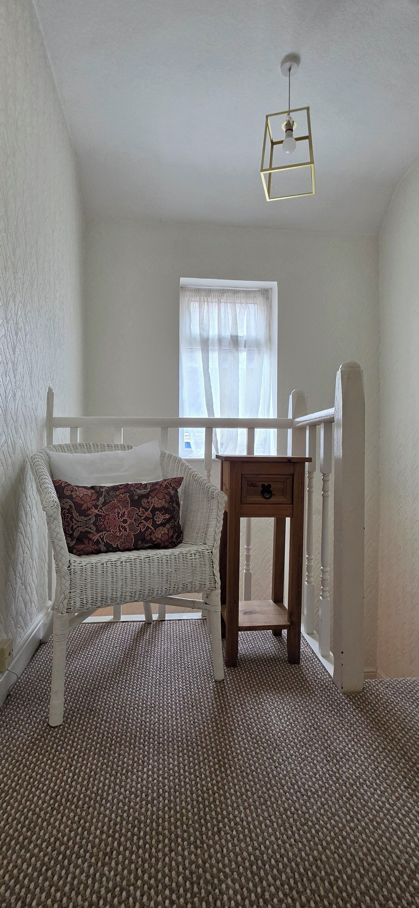 Ein Zimmer mit einem weißen Korbsessel, einem Holztisch mit Schublade und einem Fenster mit durchsichtigen Vorhängen.