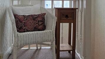 Ein Zimmer mit einem weißen Korbsessel, einem Holztisch mit Schublade und einem Fenster mit durchsichtigen Vorhängen.