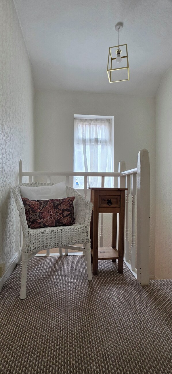 Una habitación con un sofá de mimbre blanco, una mesita de madera con cajón y una ventana con cortinas de gasa.
