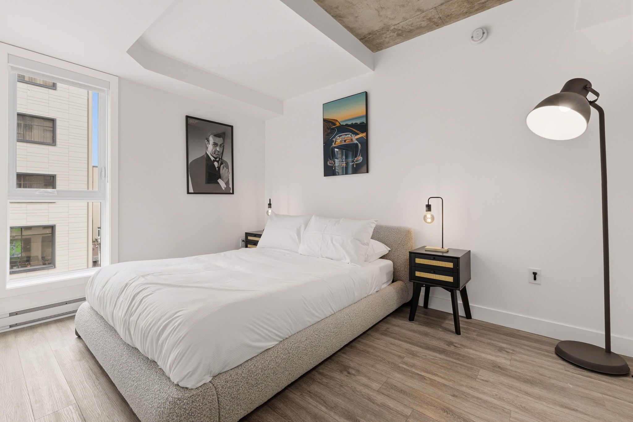A bedroom with a bed, a nightstand, a floor lamp, a window, and two wall-mounted pictures.