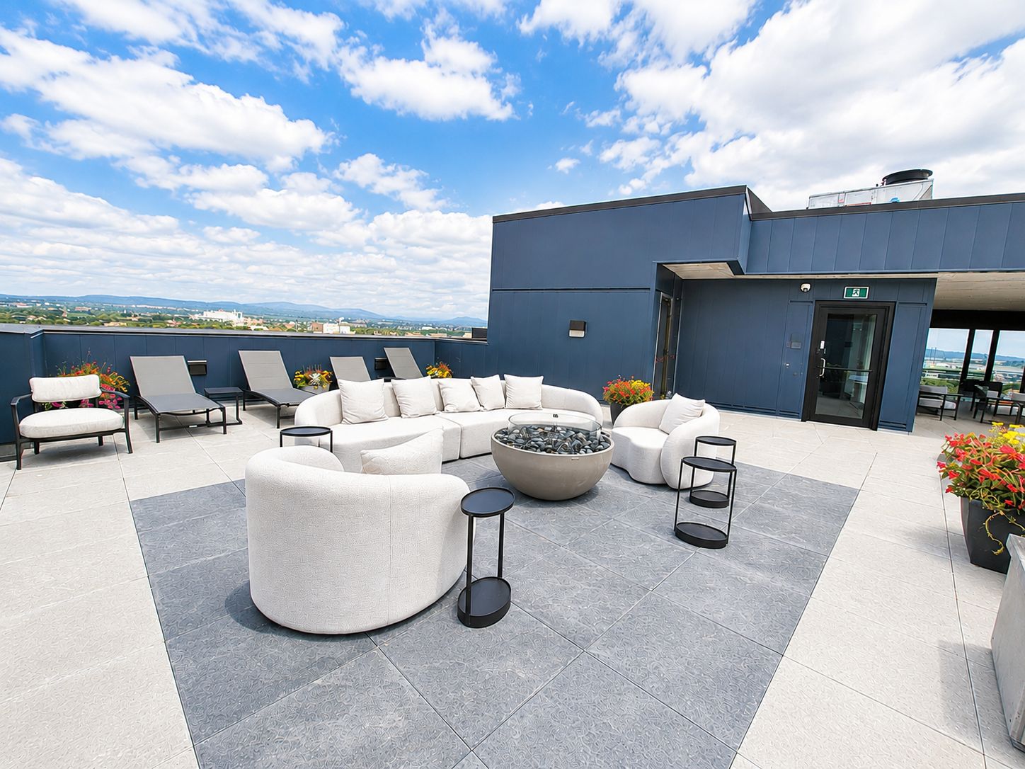 A rooftop terrace with a sectional sofa, lounge chairs, a round table, and potted plants.