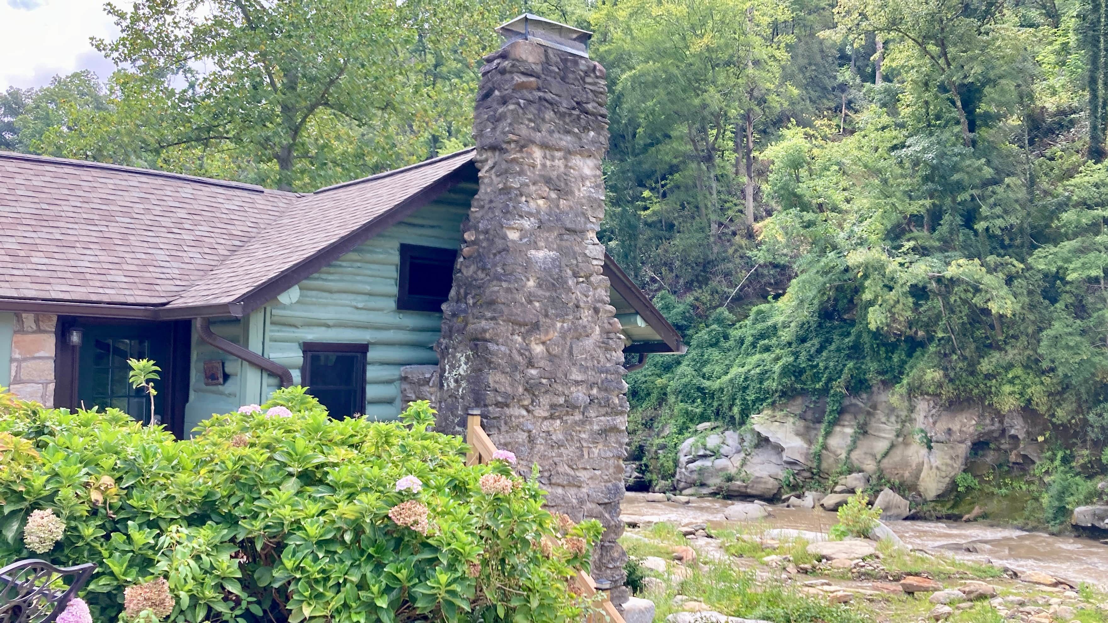A rustic cabin with a stone chimney, surrounded by lush greenery and a flowing stream.