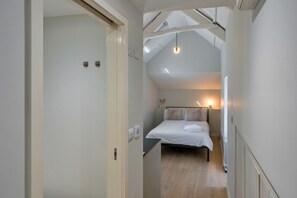A narrow hallway leading to a bedroom with a sloped ceiling, a bed with pillows, and a bedside table. - Coastal Family House for 6 (Bude)