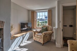 Una habitación con una televisión sobre un escritorio de madera, un sillón estampado, una ventana con contraventanas y un radiador.