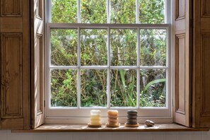 Una ventana con vista a follaje, un estante de madera con velas y una piedra en el alféizar.