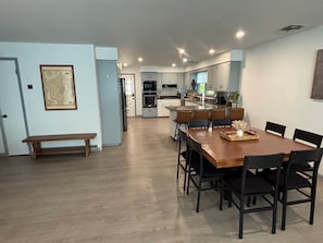Un comedor amplio con una larga mesa de madera y sillas negras, un banco, y una cocina al fondo.