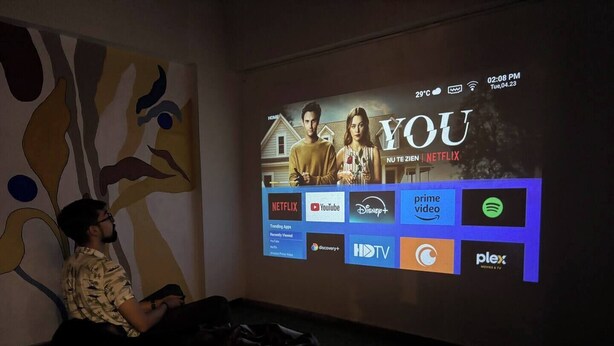 A person sitting in front of a large screen displaying a movie poster for 'YOU' on Netflix, with various streaming service logos below.
