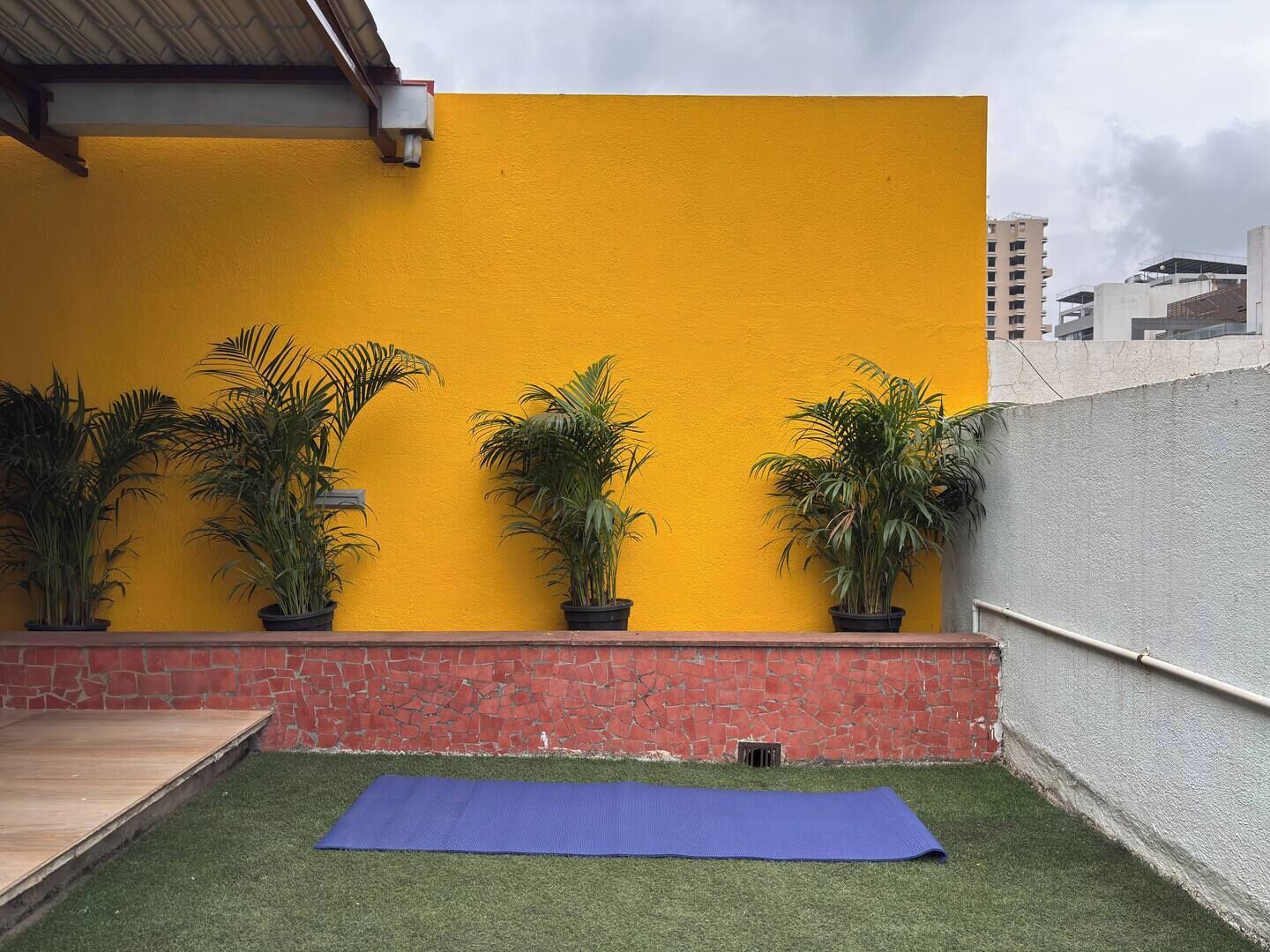 A courtyard with a yellow wall, potted plants, a blue mat, and a brick wall.