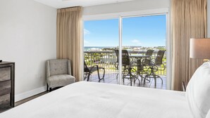 Habitación de hotel con una cama grande, una silla, una mesa con sillas y vistas al océano y edificios.
