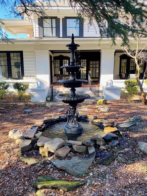 A multi-tiered fountain surrounded by rocks and foliage, with a building featuring large windows and a covered porch in the background.