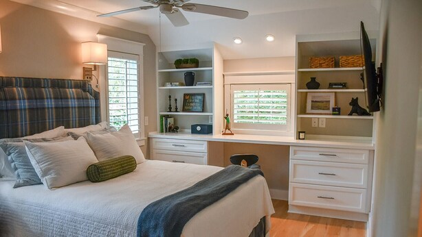 A bedroom with a bed, a desk, shelves, and a ceiling fan.