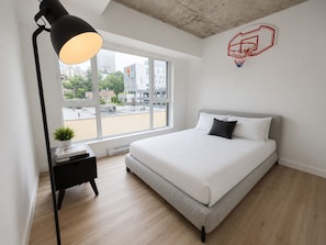 Ein modernes Schlafzimmer mit einem Bett, einem Nachttisch mit Lampe, einem Fenster mit Blick auf Gebäude und einer Wanddekoration mit einem Basketballkorb.