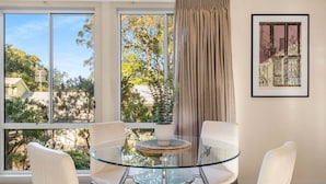 Ein Zimmer mit einem Glastisch und vier Stühlen, große Fenster mit Blick auf Bäume und Gebäude, sowie ein gerahmtes Kunstwerk an der Wand.