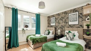 Ein Schlafzimmer mit zwei Betten, grüner Bettwäsche, einem Fenster mit Vorhängen, einem Heizkörper und Wandbildern.