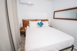A hotel room with a bed, a nightstand, a lamp, and a wall-mounted air conditioner. - Seaside Sanctuary: Bright & Airy 2BR by the Beach (Vega Alta)