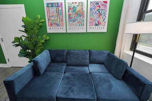 A room with a blue sectional sofa, a large green plant, and three framed artworks on the wall.