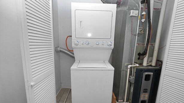 A compact, white laundry machine with control knobs and a small display, situated in a narrow space with a metal vent and pipes. - Cozy 2BR Near McCormick & Wintrust with Optional Parking (Chicago)