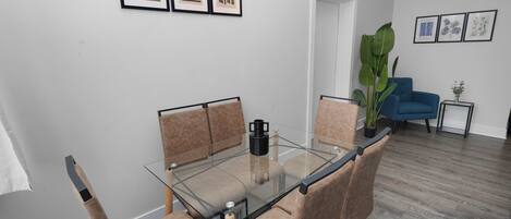 Una sala de estar moderna con una mesa de comedor de cristal, sillas con cojines color beige, un sofá azul e impresiones botánicas enmarcadas en la pared.