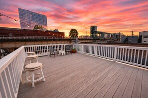 Una terraza de madera con barandillas blancas, mobiliario y vista al atardecer.