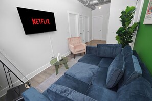 A modern living room with a blue sofa, a pink armchair, a glass coffee table, and a flat-screen TV mounted on the wall.
