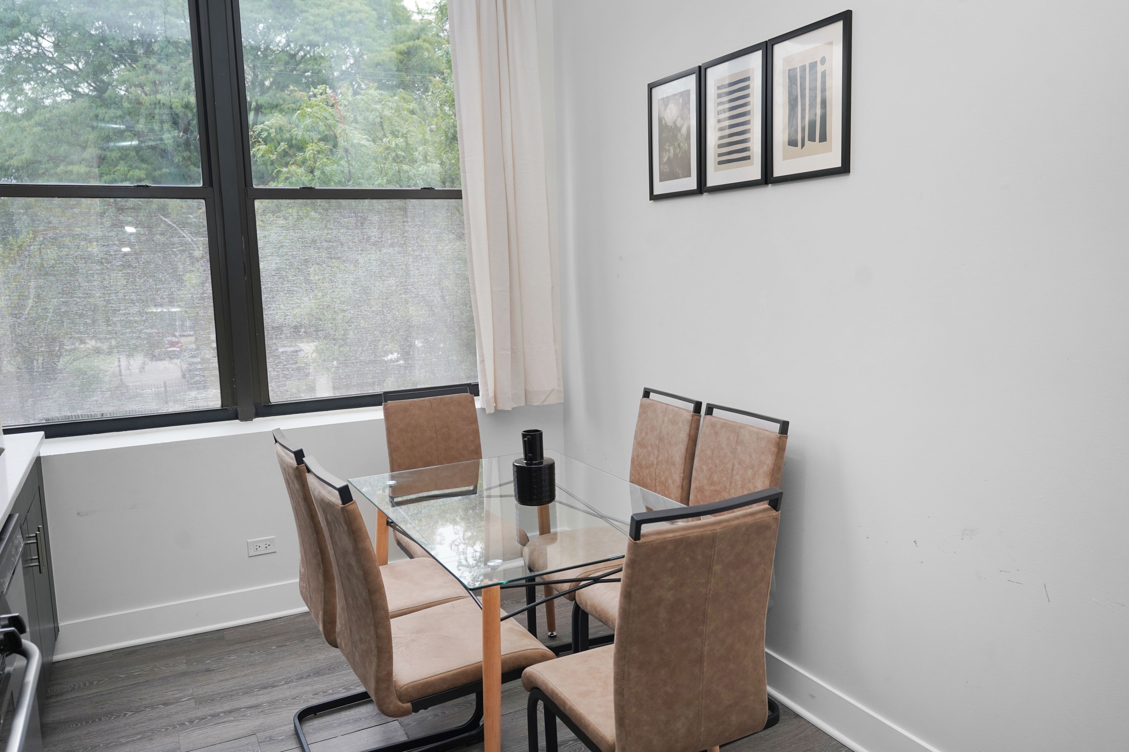 A modern meeting room with a glass table and cushioned chairs, a large window with a view of trees, and framed artwork on the wall.