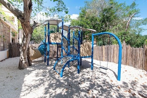 A playground with climbing structures and a slide, surrounded by trees and a wooden fence. - Elegant 2BR Condo at The Village Corasol (Playa del Carmen)