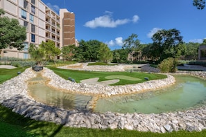 Ein angelegter Garten mit Teich, umgeben von einer Steingrenze, und ein Gebäude mit mehreren Balkonen im Hintergrund.