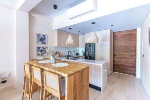 A modern kitchen with a central island, wooden chairs, a hanging light fixture, and a large wooden door. - 2BR Resort Condo near Beach & Golf (Playa del Carmen)
