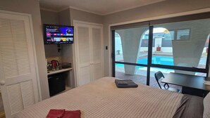 A hotel room with a bed, a TV on the wall, a small kitchenette, and a view of a swimming pool.