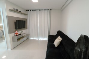 A modern living room with a black sofa, a flat-screen TV mounted on the wall, a small table with a plant, and a window with white curtains. - Family Apartment III near Beto Carrero World and the Beach (Penha)