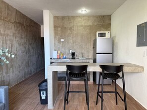 Ett kompakt kök med ett kylskåp, en mikrovågsugn och en liten arbetsbänk. Det finns två svarta barstolar och en papperskorg.