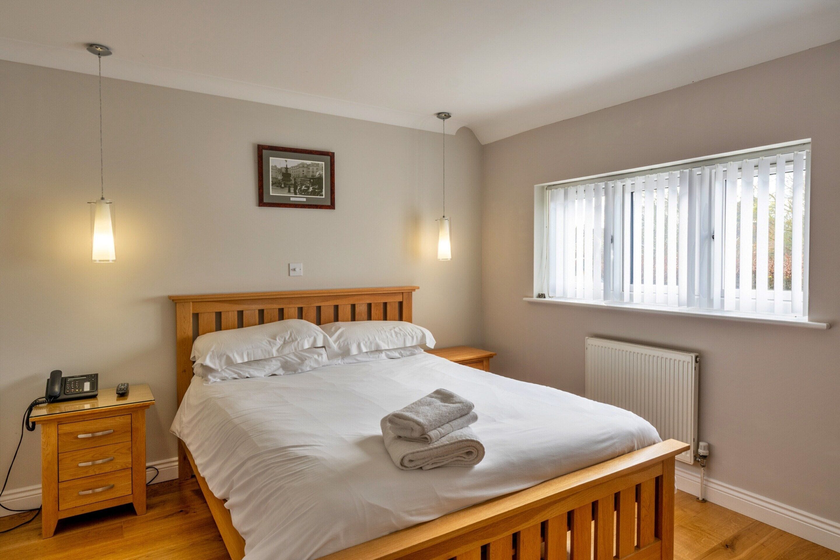 A bedroom with a wooden bed, bedside tables, a telephone, a framed picture on the wall, and a window with blinds.