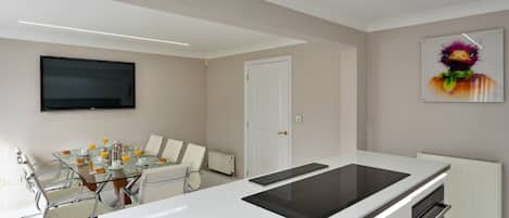 A modern kitchen with a central island, a dining area with a glass table, and a flat-screen TV mounted on the wall.